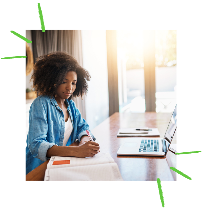 A young mother with coily hair wearing a blue denim button down shirt and a white shirt underneath is sitting at a desk infront of a laptop. She is taking notes in a notebook while receiving coaching session for her child's upcoming IEP Meeting